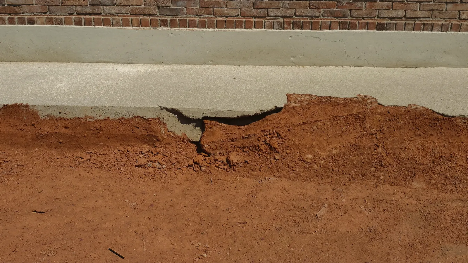 Exposed edge of a concrete slab-on-grade foundation where the dry red clay has pulled away, leaving a visible gap beneath the slab perimeter