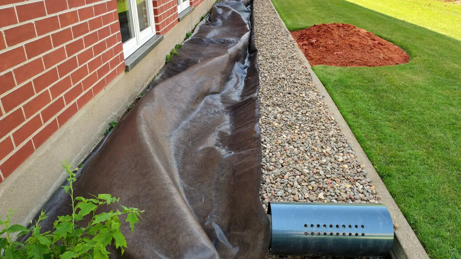 Open french drain trench lined with filter fabric and washed gravel around perforated pipe, running along the side yard of a brick house
