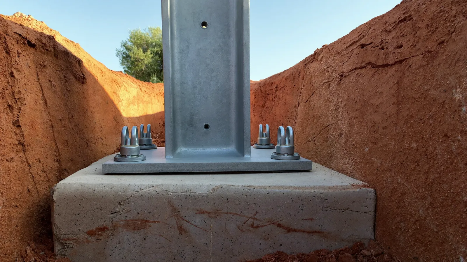 Close-up of a galvanised steel push pier bracket bolted to a concrete footing in a narrow excavation, pier sections stacked in the red clay beside it